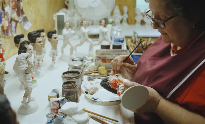 A lady paints a Grogg with red paint, more unfinished Groggs sit on the table.